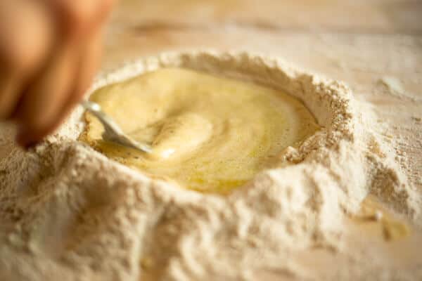 a fork mixing liquid in a flour mound on a wooden surface
