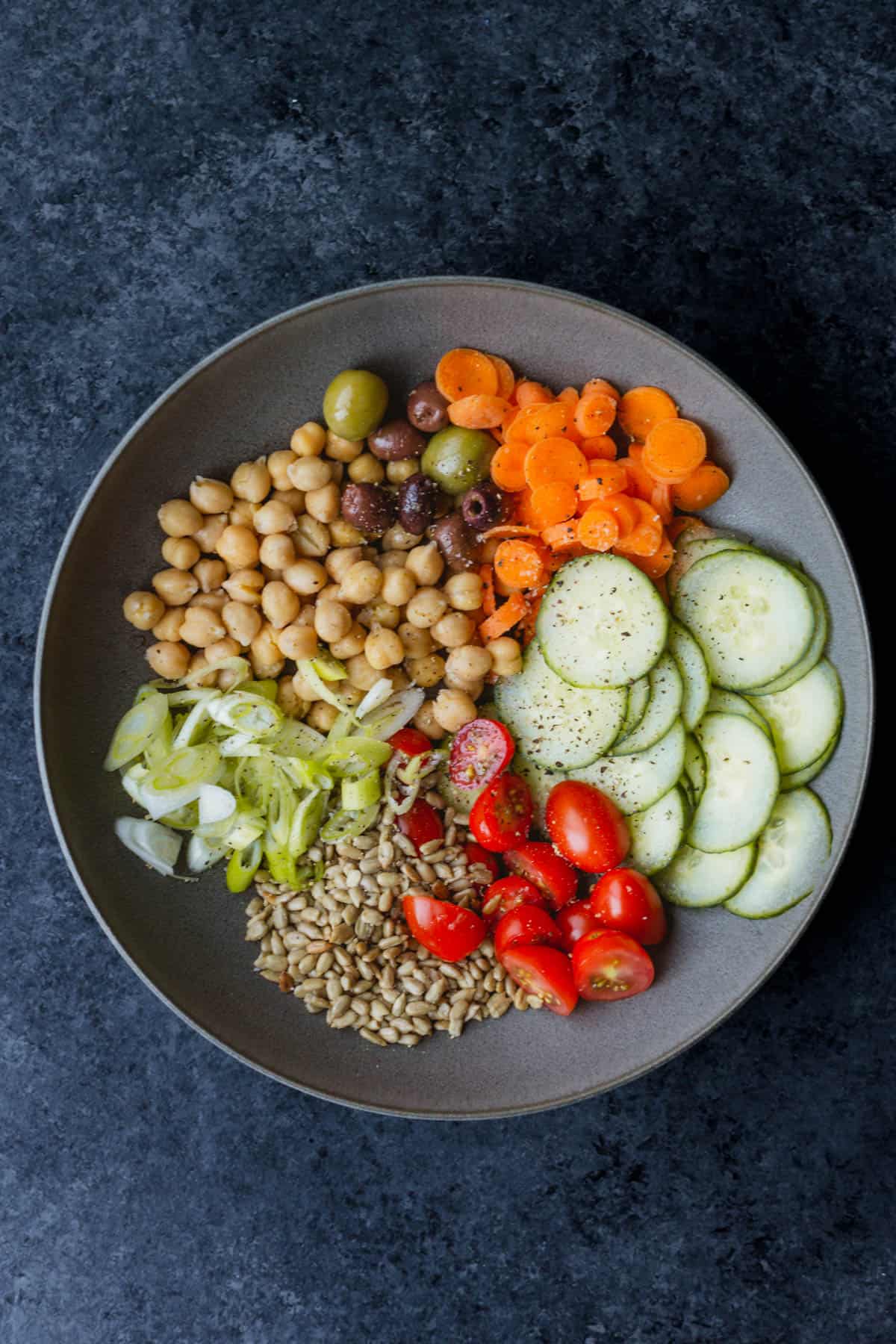 a lettuceless salad of cucumbers, tomatoes, sunflower seeds, green onions, chickpeas, olives, and carrots in a dark grey bowl