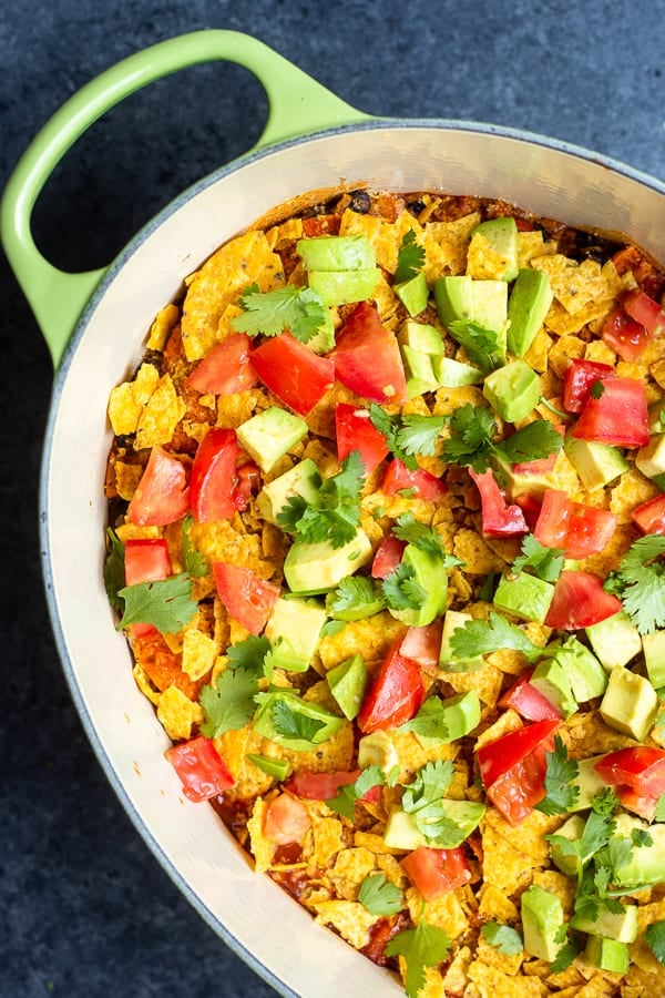 an oval dutch oven filled with a one-pot vegan enchilada casserole and topped with avocado, chips, tomato, and cilantro
