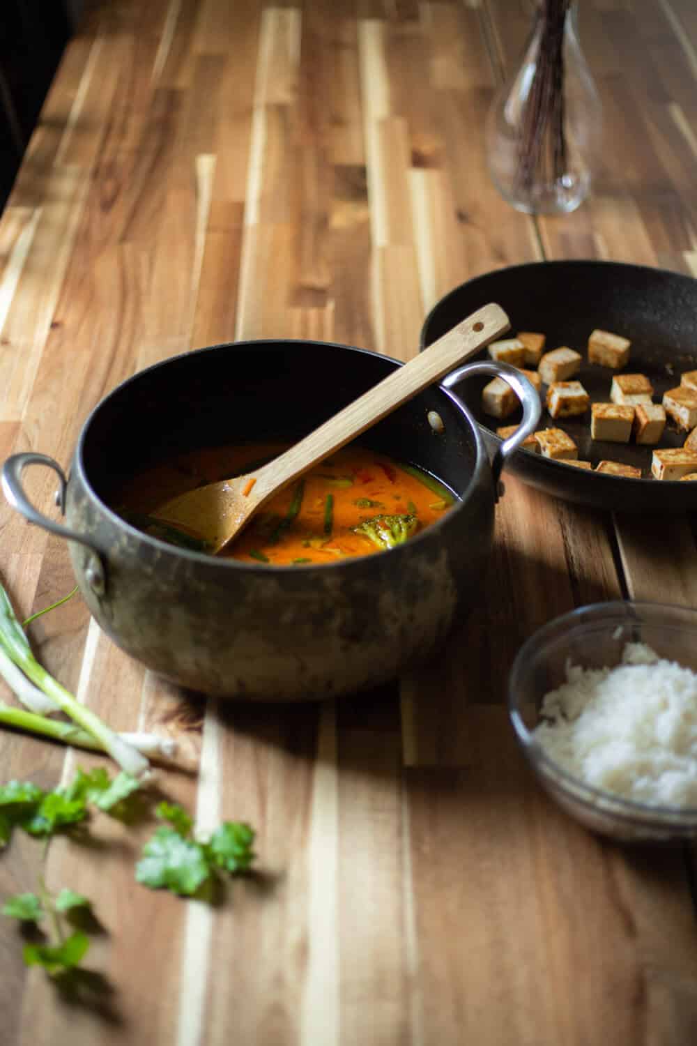 vegan pineapple curry in a pot with a pan of crispy tofu, a bowl of rice, and green onions and cilantro surrounding it