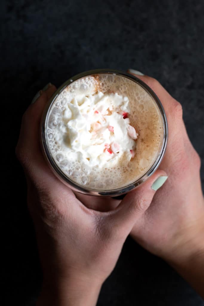 a girl's hands wrapped around a vegan oat milk peppermint mocha latte in a glass cup